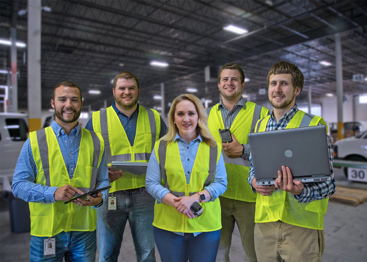 Employees in warehouse holding various forms of technology such as laptops, mobile phone, thumb scanner and tablet